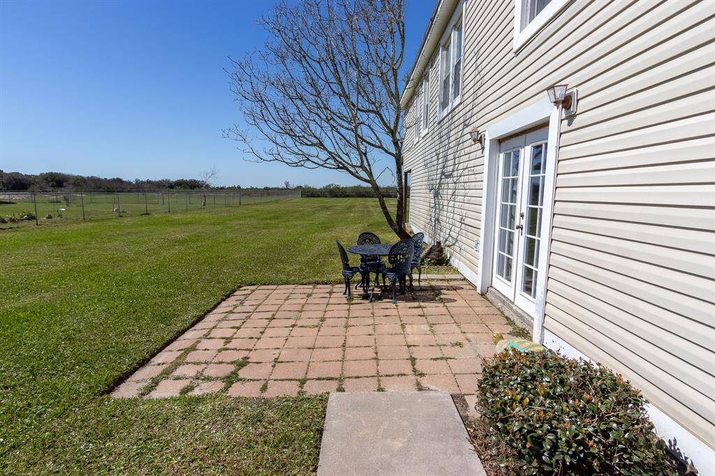 This photo shows the spacious yard with a patio area. The house has light siding and French doors leading outside, offering a view of an expansive, fenced green field, perfect for outdoor activities.