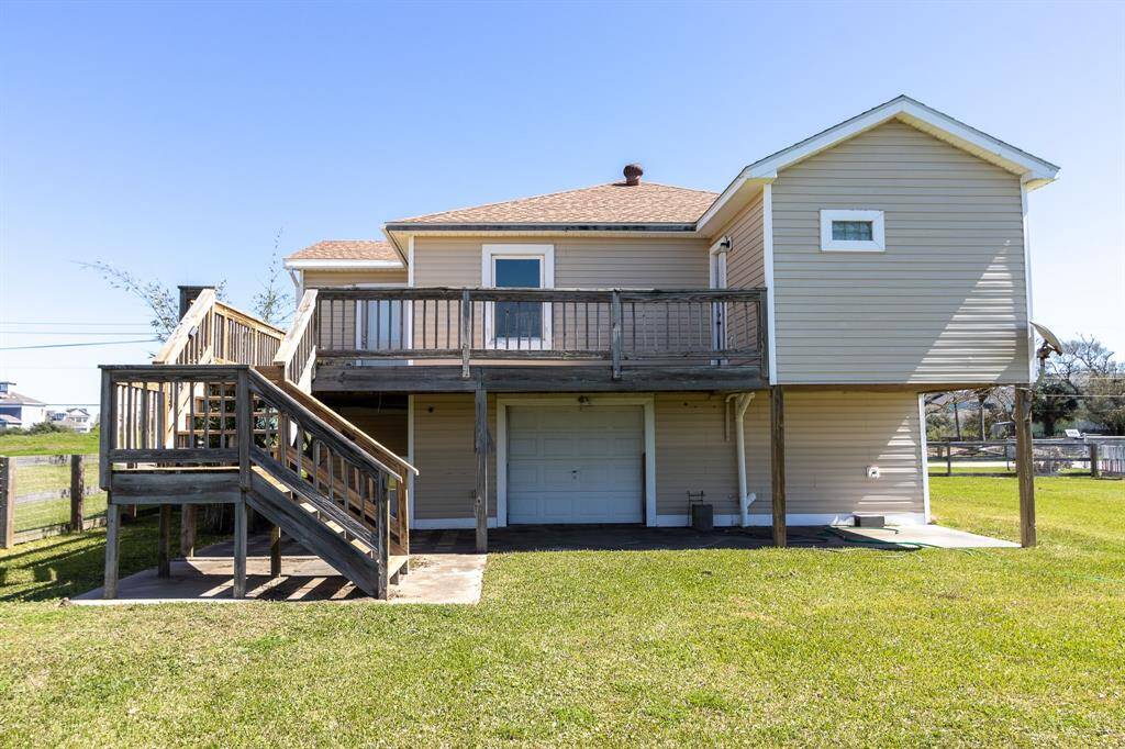 This photo shows the back side of the home. It has a wooden staircase leading to a roomy balcony and a covered area beneath, complete with a garage door. The yard is spacious and grassy, ideal for outdoor activities.