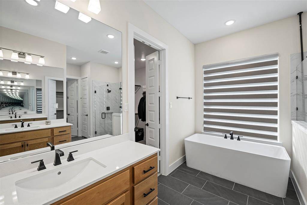 A deep soaking tub sits beneath a large window with modern dual shades, offering privacy and natural light. The layout seamlessly blends luxury and function, complete with a spacious walk-in closet just beyond the bath.