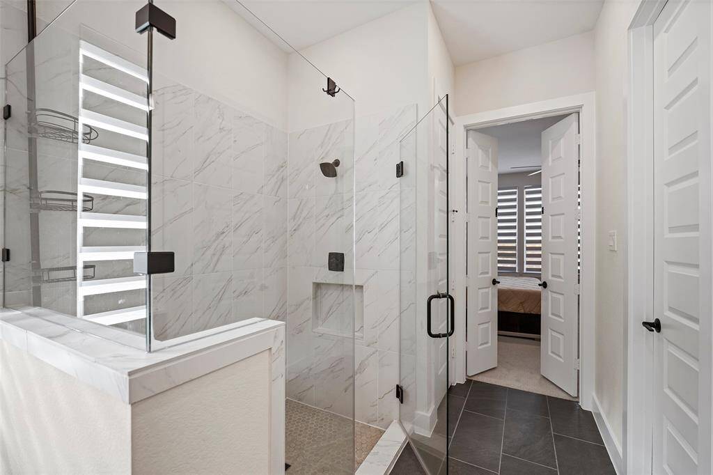 Frameless glass surrounds the walk-in shower, showcasing marble-look tile and matte black fixtures for a clean, modern aesthetic. Built-in shelving offers practical storage while maintaining a streamlined design.