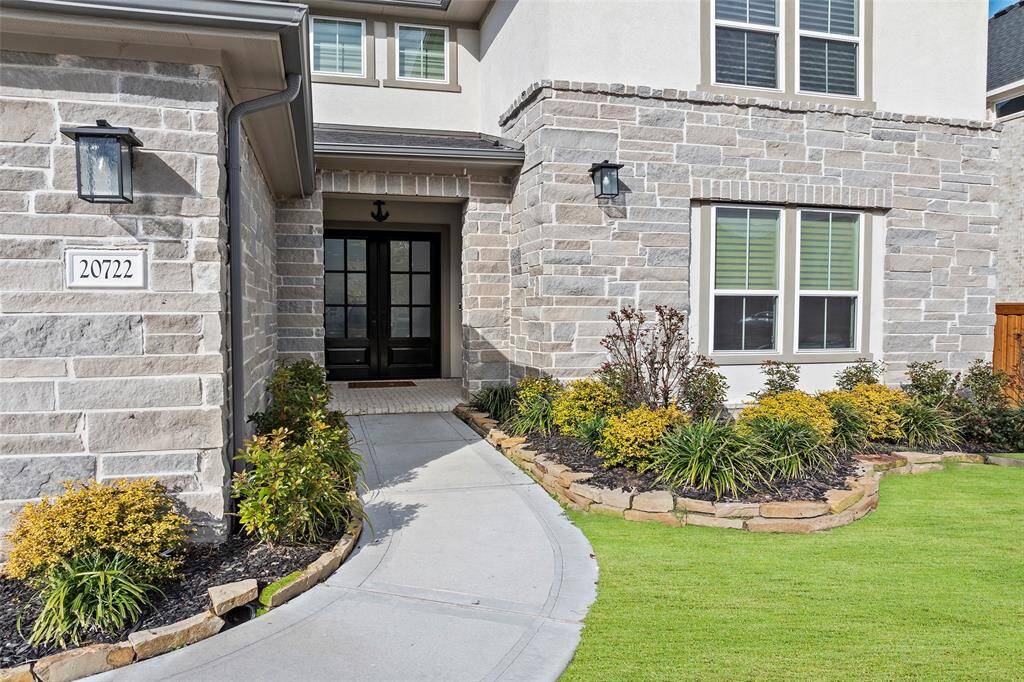 A winding walkway bordered by stone-lined garden beds leads to a set of striking double doors, framed by a stone and stucco exterior. Fresh landscaping and thoughtful architectural details add charm and curb appeal, setting a warm and welcoming tone before you even step inside.