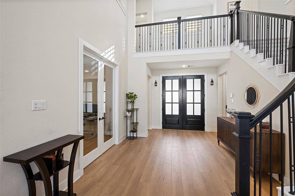 A dramatic two-story foyer welcomes you with a sweeping staircase, black double doors, and warm wood flooring that flows throughout the home. Natural light pours in from above, highlighting the sleek iron railing and creating a grand yet inviting first impression. A set of French doors opens to a flexible space—perfect for a study, playroom, or media room.