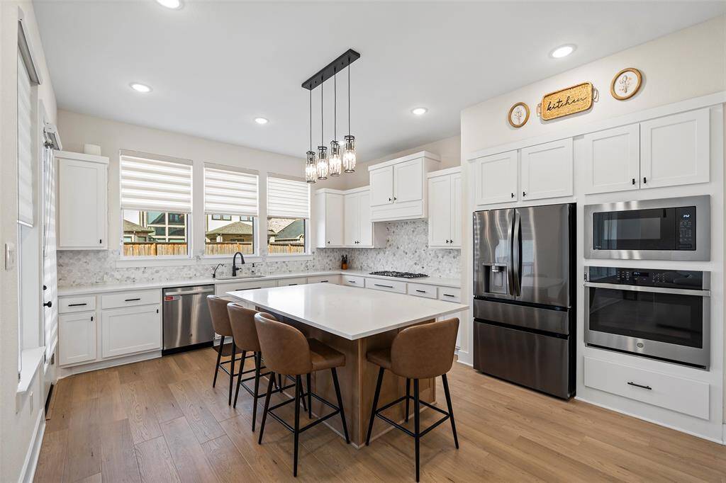 Crisp white cabinetry, quartz countertops, and stainless steel appliances anchor this thoughtfully designed kitchen. A large center island with bar seating creates the perfect setting for casual dining or entertaining, while windows along the back wall flood the space with natural light.