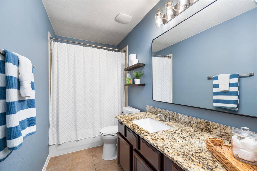 This beautifully maintained bathroom exudes timeless elegance with its granite countertop, modern sink, and stylish lighting. The blue walls and tiled floor add a serene touch, while the spacious shower with a simple white curtain enhances the clean, contemporary look. Wooden shelves provide functional storage, perfectly complementing the room’s refined design.