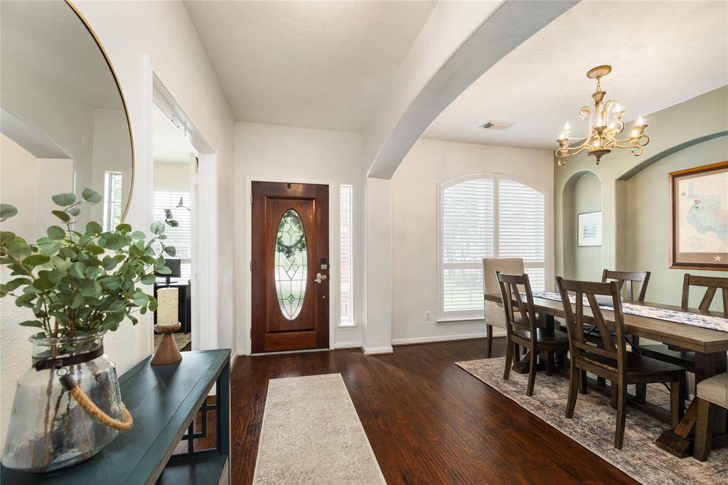 Step into an inviting entryway where a beautifully crafted front door opens into a bright and elegant dining room. Arched doorways, a graceful chandelier, and rich wood flooring set the tone for timeless sophistication, while large windows, a statement mirror, and tastefully styled accents complete the welcoming ambiance.