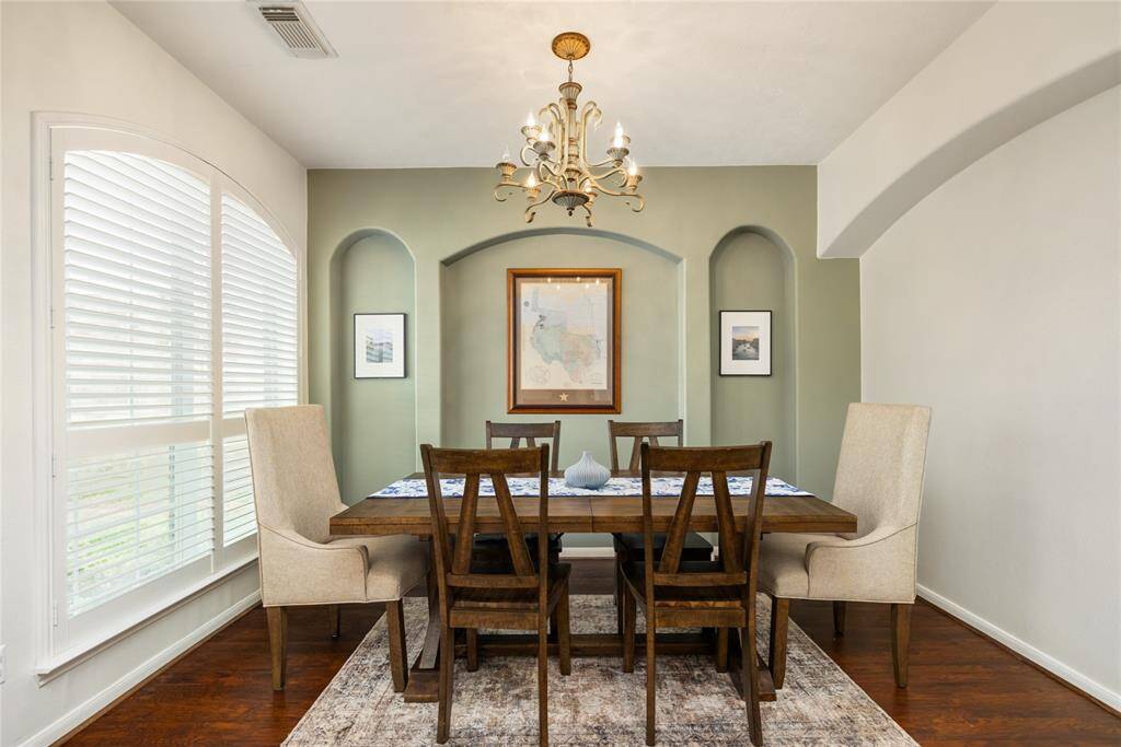 This formal dining room exudes timeless sophistication with its arched wall niches, elegant chandelier, and soft natural light streaming through plantation shutters. Rich hardwood flooring anchors the space, offering the perfect setting for memorable gatherings in a home that’s been thoughtfully maintained and beautifully presented throughout.