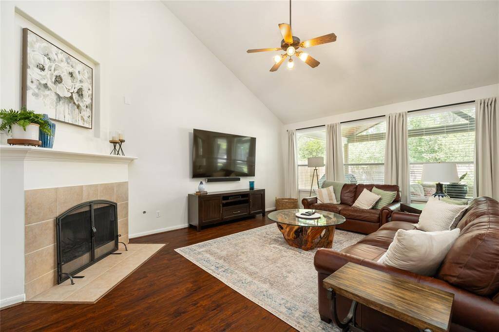 This spacious living room features a vaulted ceiling with a ceiling fan, a cozy fireplace, and large windows for natural light. It has hardwood floors, and comfortable leather seating, creating a warm and inviting atmosphere.