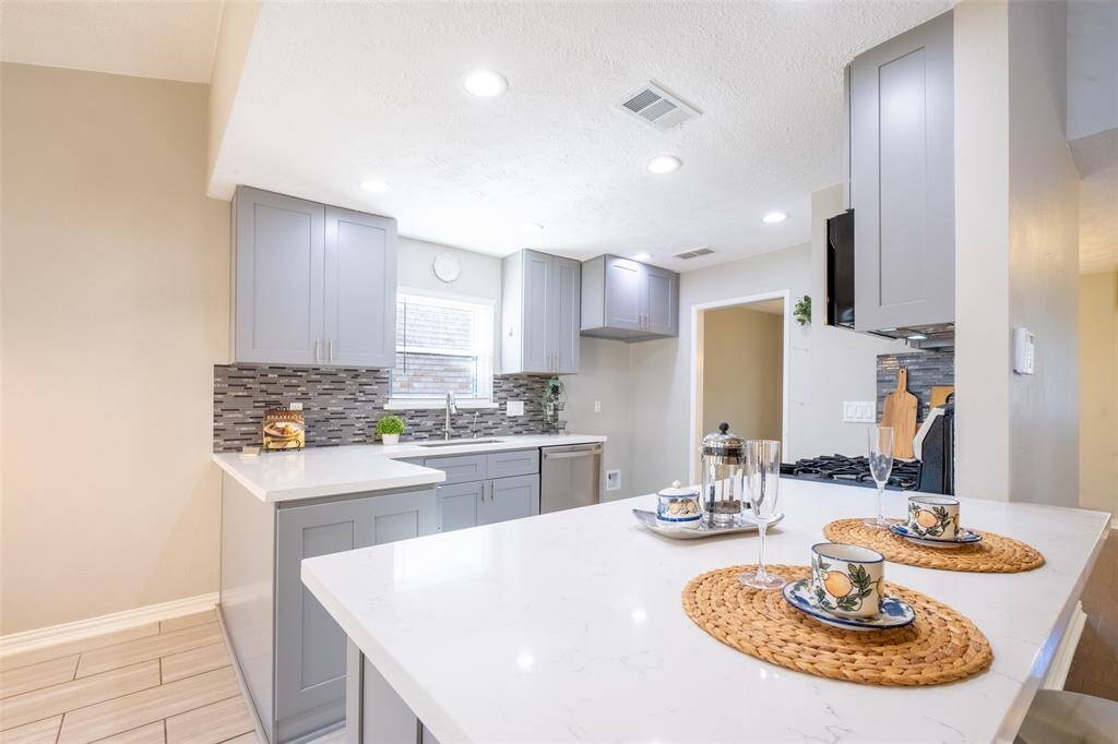 This modern kitchen features sleek gray cabinets, white countertops, and a stylish tile backsplash. It includes a spacious island with seating, perfect for casual dining. Recessed lighting adds brightness, while a window above the sink provides natural light.