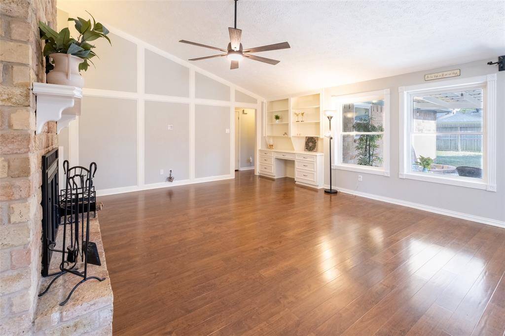 Bright, spacious living room featuring hardwood floors, a brick fireplace, built-in shelving, and large windows overlooking the backyard. High ceilings and neutral colors enhance the airy feel.