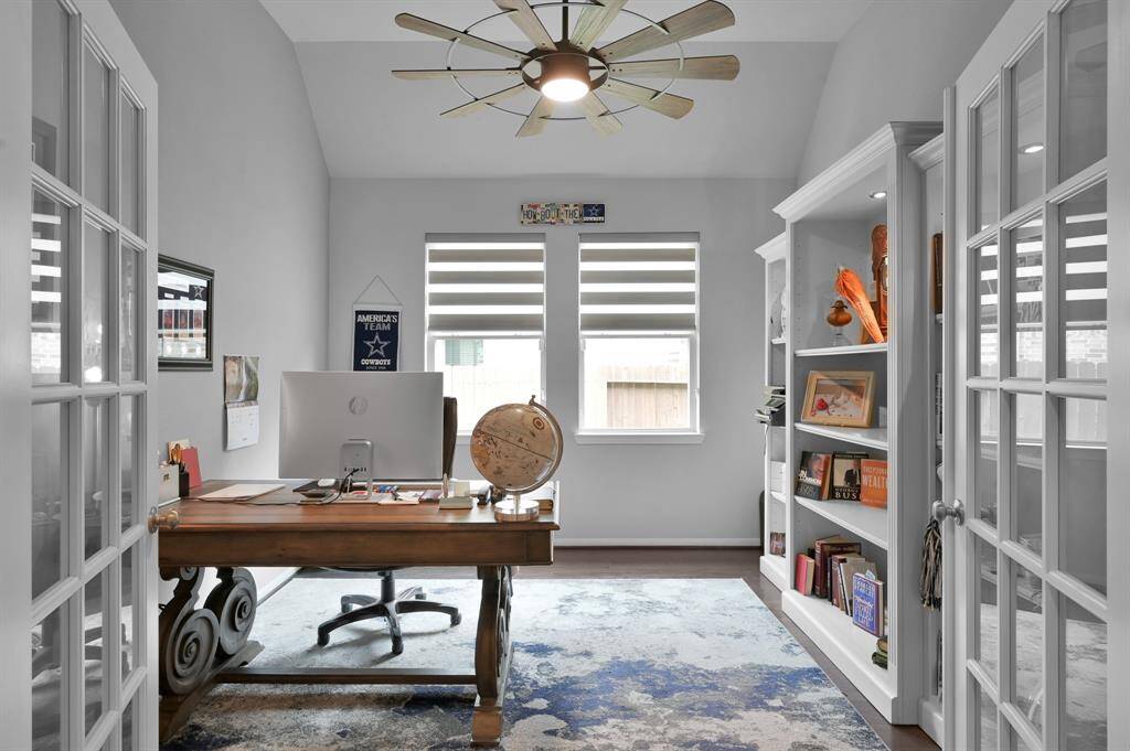 This photo showcases a bright home office with glass French doors, built-in bookshelves, and a modern ceiling fan. The room has a cozy, organized feel with ample natural light from the window.