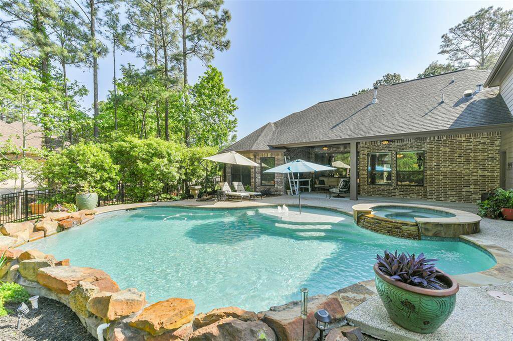 A tranquil backyard oasis with a free-form pool and attached circular spa, framed by natural stone coping and a pebble-finish deck. A single large umbrella shades the shallow sun shelf, while cushioned chaise lounges sit under a second umbrella nearby. A wrought-iron fence and mature pines provide privacy and a lush, forest-like backdrop.