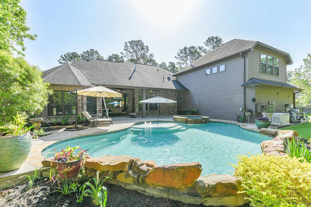 This image showcases a beautifully landscaped backyard with a sparkling private pool. The pool features a built-in spa, small water fountain jets, and is surrounded by natural stone accents. The home exterior combines brick and siding, with a covered patio area that offers a relaxing shaded space. Lush greenery, potted plants, and well-maintained landscaping add to the serene and inviting atmosphere. Tall trees in the background provide additional privacy and a peaceful natural backdrop.