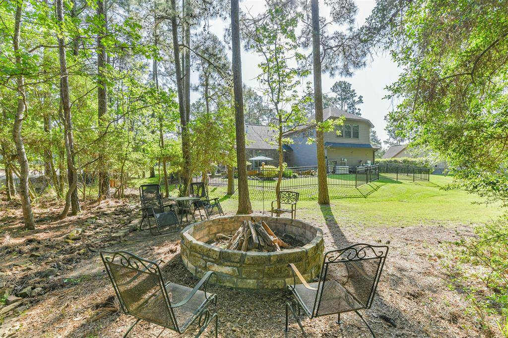 This image features a peaceful wooded backyard retreat with a rustic stone fire pit as the centerpiece. Surrounded by tall pine trees and natural landscaping, the space feels private and serene. In the distance, you can see a well-maintained lawn and the main house, which includes a fenced-in pool area. The blend of natural shade and open space creates a relaxing atmosphere perfect for quiet evenings or outdoor gatherings.