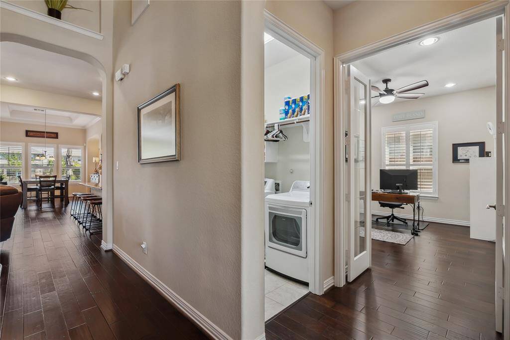 Hallway view of an office space, with a view of laundry room and open views of the dining room, kitchen, and den. French doors are in the foreground. Also view is a space in the laundry room where a refrigerator can be placed.