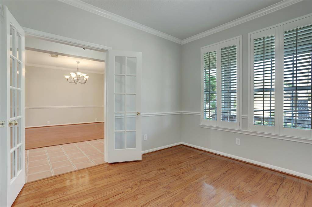 This photo showcases the office with hardwood flooring, large windows with shutters, and elegant French doors leading to another area with a chandelier. The walls are painted in a neutral tone, and there's decorative molding for a classic touch.
