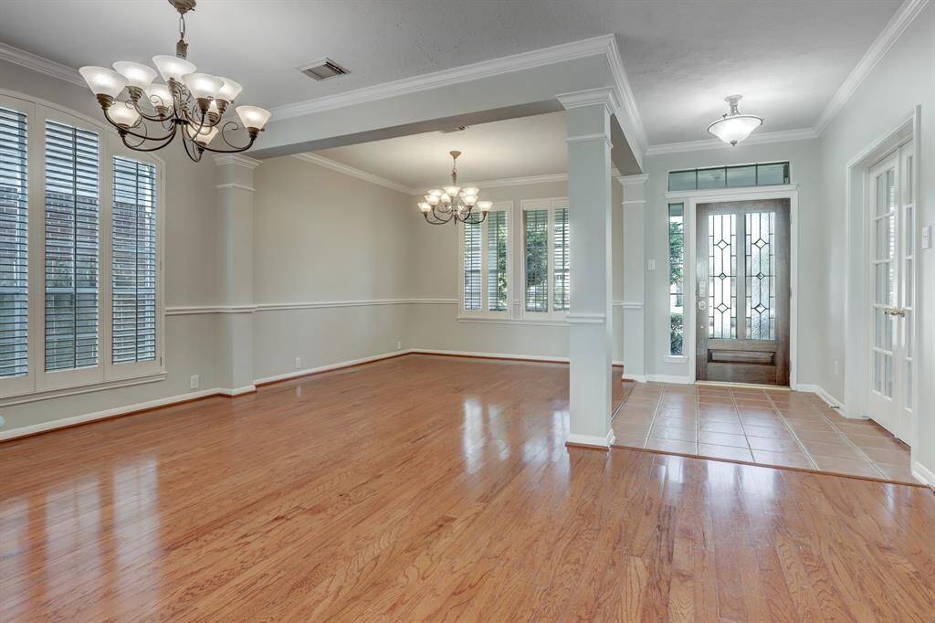 Bright and spacious open-concept living area with hardwood floors, elegant chandeliers, and large windows with shutters. The entryway features decorative glass doors and tiled flooring, enhancing the welcoming atmosphere.