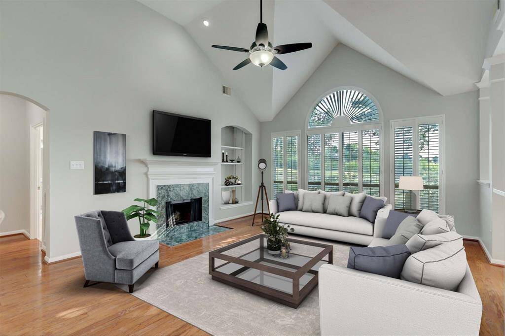 This spacious living room features high vaulted ceilings, a large arched window with plantation shutters, and a cozy fireplace. It’s complemented by elegant hardwood floors.