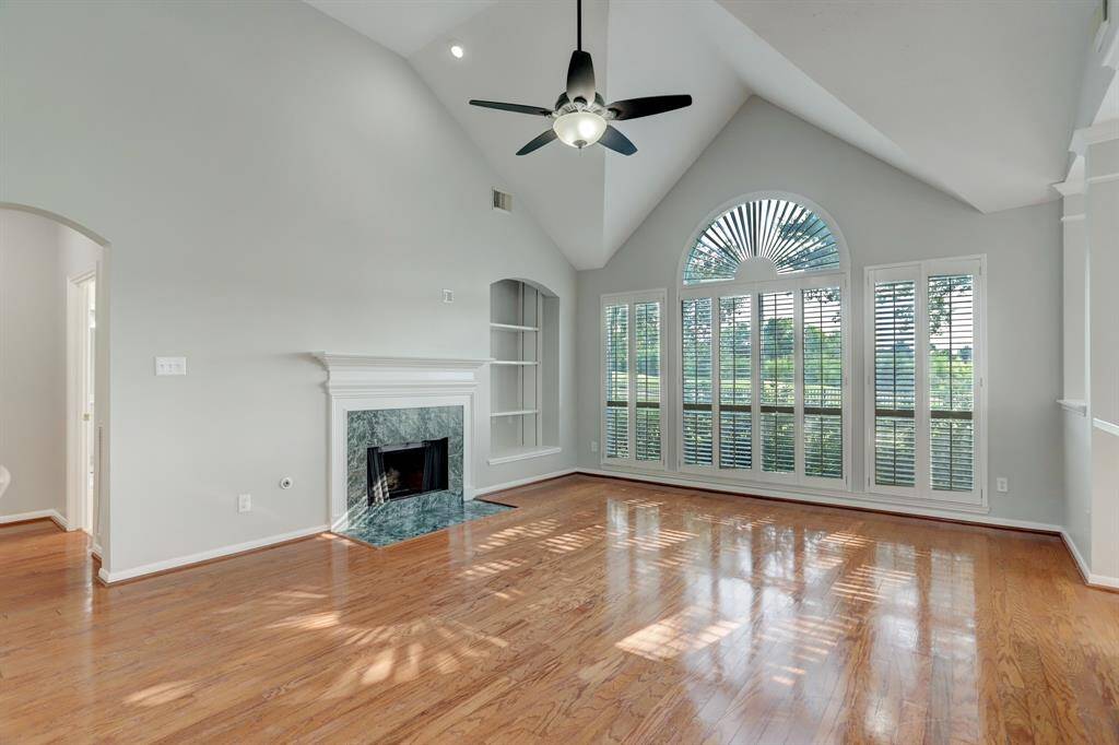 Bright and spacious living room with high ceilings and a ceiling fan. Features a large arched window with plantation shutters, a cozy fireplace, and elegant hardwood floors. Ideal for natural light and comfort.