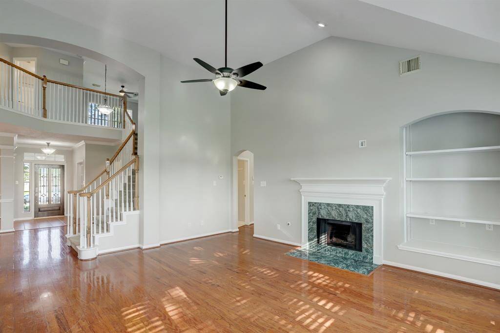 This spacious living area features high ceilings, a modern ceiling fan, and hardwood floors. It includes a fireplace with a marble surround, built-in shelving, and an elegant staircase leading to an upper level. Abundant natural light enhances the open, airy feel.