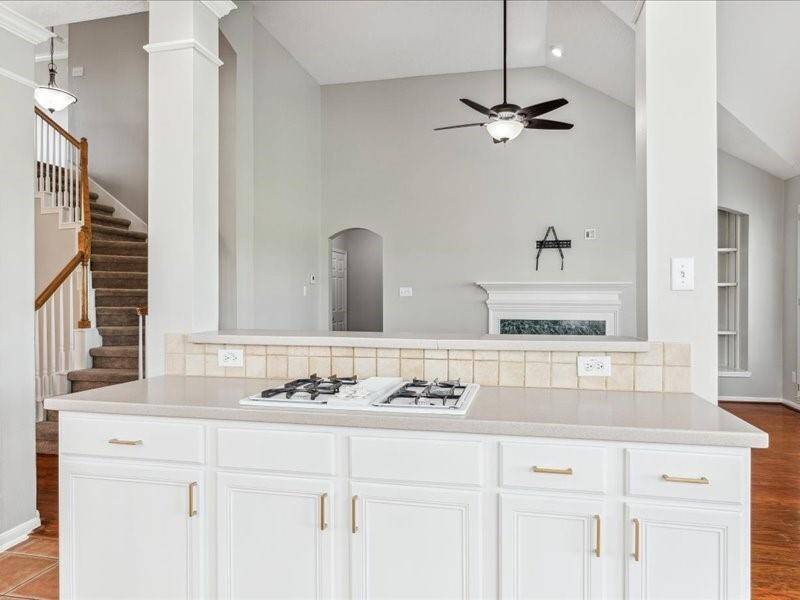 This photo showcases a bright, open living area with a modern kitchen featuring a gas cooktop on a large island. The space includes high ceilings, a ceiling fan, a cozy fireplace, built-in shelving, and a staircase, all with a neutral color palette.