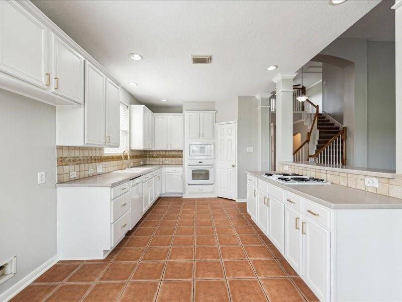 This spacious kitchen features white cabinetry, tile flooring, and ample counter space, with built-in appliances and a view of the staircase, creating a bright and open atmosphere.