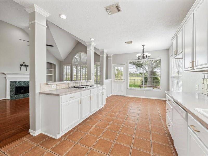 This bright kitchen features white cabinetry, a spacious island with a cooktop, and terracotta tile flooring. It opens to a living area with hardwood floors and a fireplace, and has large windows that offer views of the backyard.