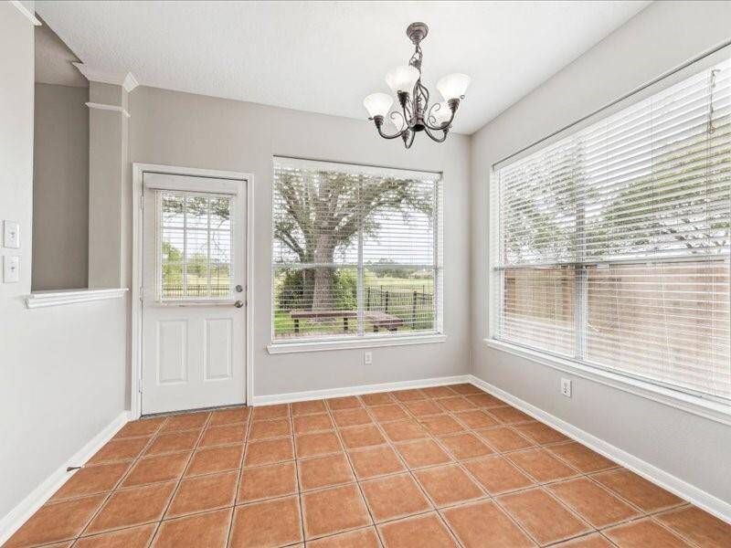 This bright room features terracotta floor tiles, large windows with white blinds offering a view of a tree-filled backyard, and a stylish chandelier. It includes a door leading outside, perfect for a cozy dining area or sunroom.