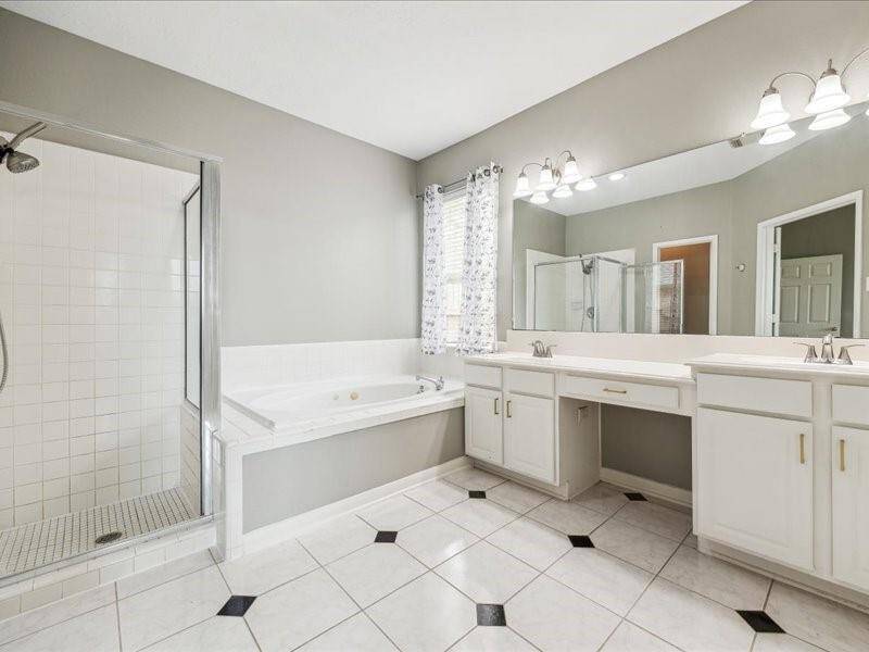 This spacious bathroom features a glass-enclosed shower and a luxurious corner bathtub. It has a large double vanity with ample storage, bright lighting, and neutral tile flooring with decorative accents.