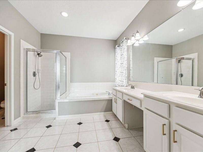 This spacious bathroom features a modern design with neutral tones, a large dual-sink vanity, a soaking tub, and a separate glass-enclosed shower. The room is well-lit with recessed lighting and has tile flooring with decorative accents.