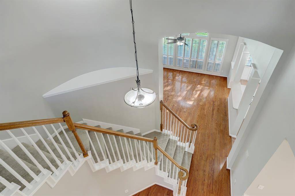 This photo features a bright, open stairway with wooden handrails and white balusters, leading to a spacious room with hardwood floors and large windows with shutters, offering ample natural light.