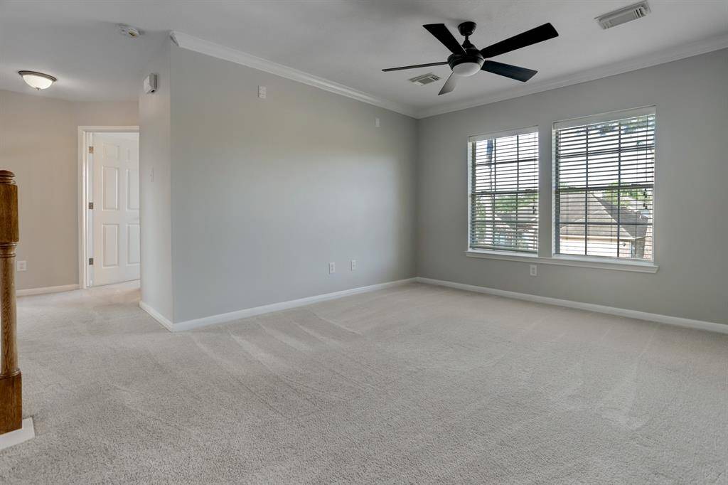 This spacious, light-filled room features neutral carpeting, soft gray walls, and a ceiling fan. Large windows provide ample natural light, creating a welcoming atmosphere.