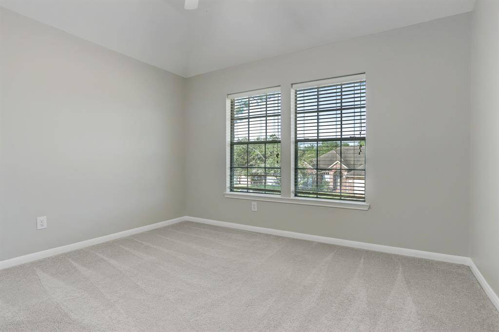 This is a bright and spacious room with light gray walls and carpet. It features two large windows with blinds, offering a pleasant view and ample natural light.