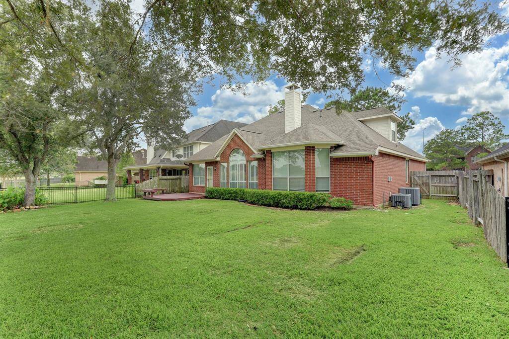 This photo shows a spacious backyard with a well-maintained lawn and mature trees, featuring a red-brick house with large windows and a chimney. There is a wooden deck on the side, ideal for outdoor gatherings, and a fenced perimeter for privacy.