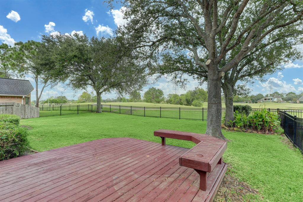 Spacious backyard featuring a wooden deck with built-in seating, lush green lawn, and mature trees, enclosed by a black metal fence, perfect for outdoor relaxation and entertaining.