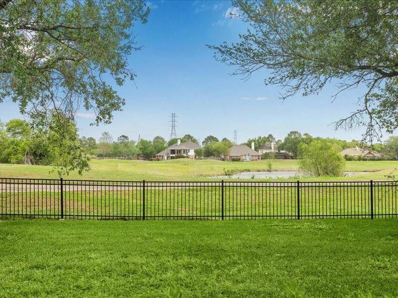 This photo showcases a spacious backyard with a well-maintained lawn and a black metal fence. It offers a picturesque view of a serene landscape with a pond and neighboring homes in the distance, framed by mature trees under a clear blue sky.