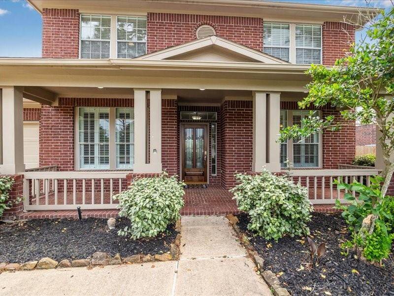 This charming two-story home features a welcoming brick exterior with a covered front porch, accented by white railings and lush landscaping. The pathway leads to a stylish front door, offering an inviting entrance.