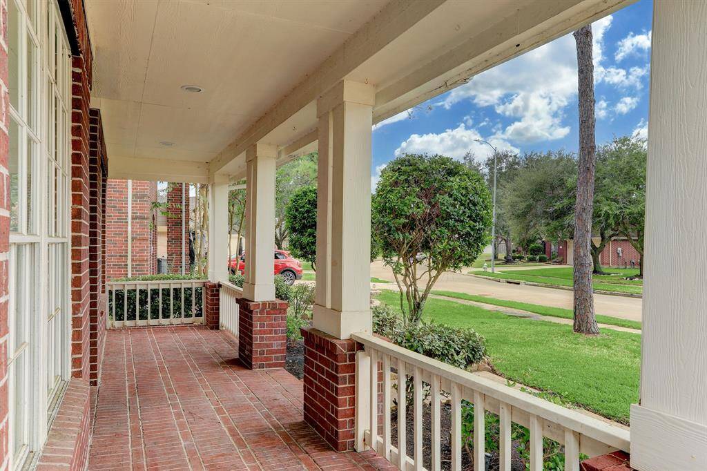 Charming front porch with red brick flooring and columns, offering a cozy view of a tree-lined street. Ideal for relaxing outdoors.
