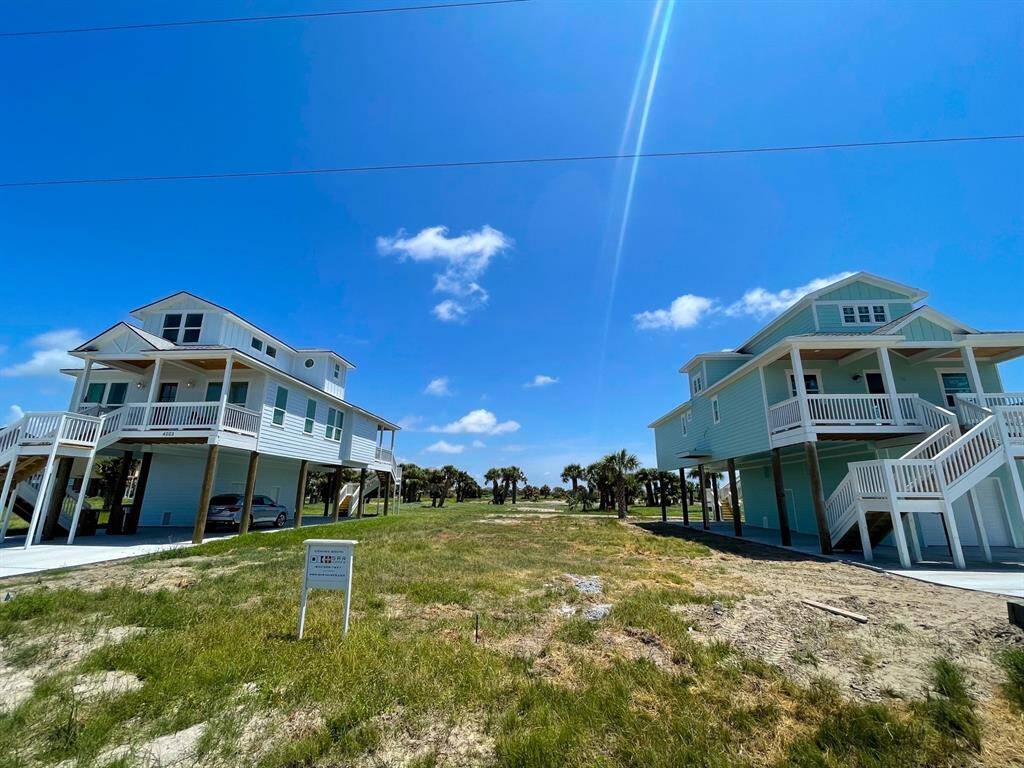 This photo shows two elevated coastal homes with large front porches, set on a spacious lot with grassy surroundings. The sky is clear and sunny, creating a bright and inviting atmosphere.
