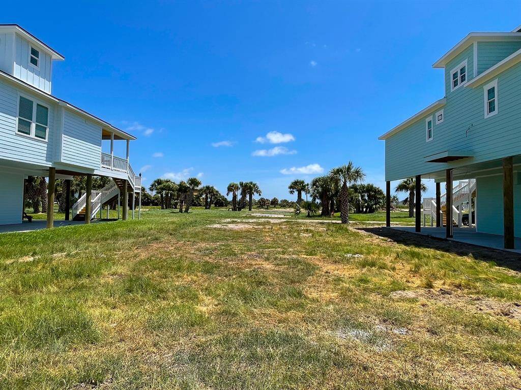 This photo shows a coastal property with elevated houses on either side, set against a backdrop of palm trees and a bright blue sky. The area is grassy and open, suggesting a serene, beachside setting.