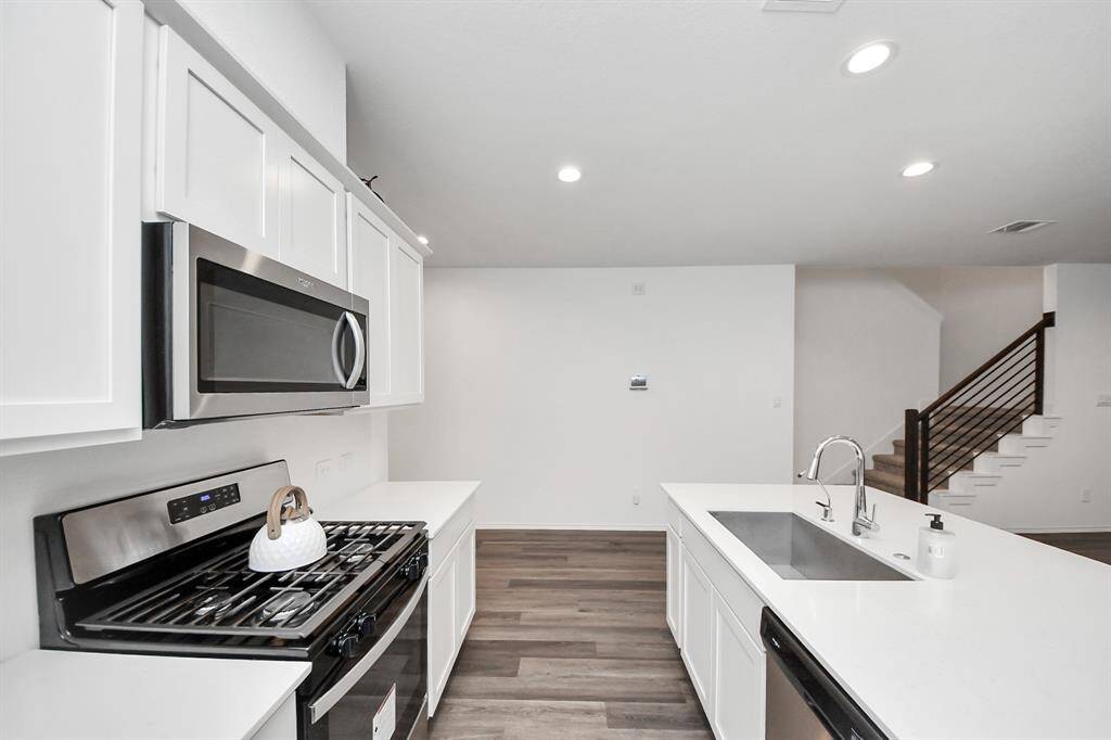 This modern kitchen features sleek white cabinetry, stainless steel appliances, and a spacious island with a sink. It opens to a bright living area with recessed lighting and a staircase with stylish railing, enhancing the open-concept design.