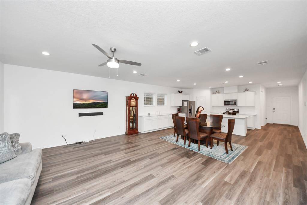 This open-concept living space features modern wood laminate flooring, a ceiling fan, and recessed lighting. The room includes a cozy sitting area, a dining table, and a sleek, white kitchen with stainless steel appliances. A wall-mounted TV and a classic grandfather clock add charm to the space.