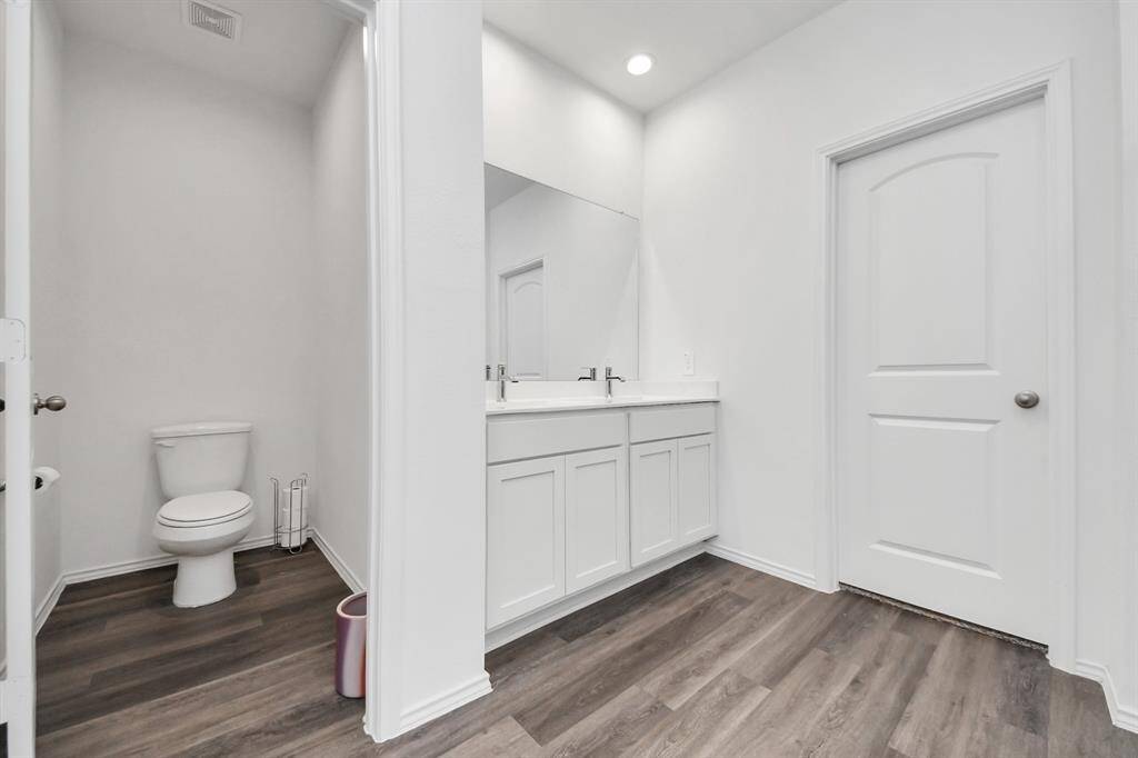 a modern bathroom with a dual-sink vanity and large mirror. It features white cabinetry, recessed lighting, and wood-look flooring. There is a separate toilet area for added privacy.