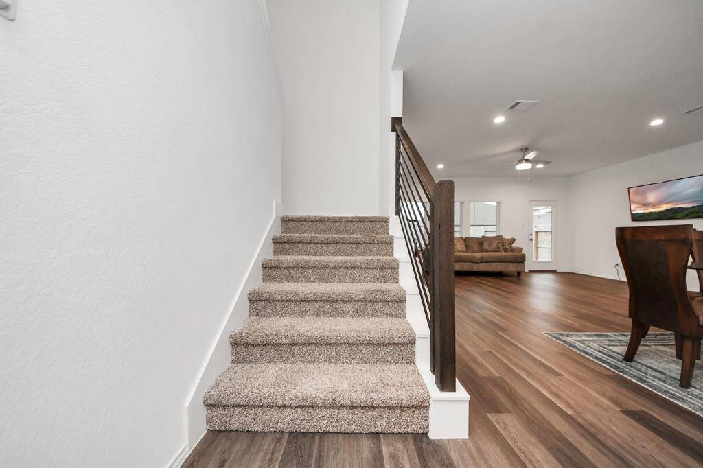 a modern, open-concept living area with wood flooring and a carpeted staircase. It features a cozy living room with a ceiling fan, adjacent dining space, and ample natural light from large windows and a glass door.