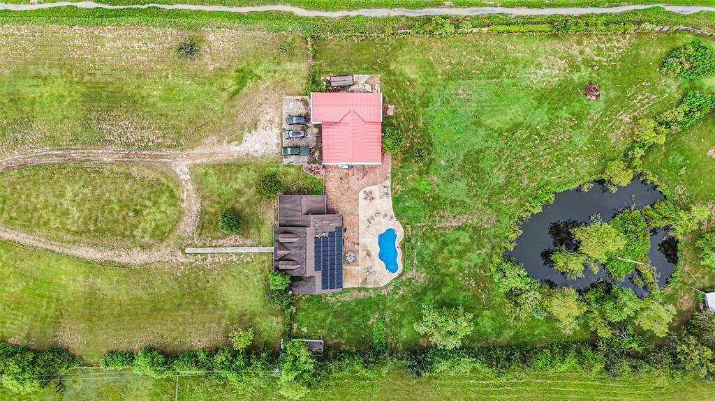 This aerial image showcases a rural property with a spacious lot, featuring a large shop with a red roof. The property includes a private pond and a swimming pool, offering a blend of natural and landscaped outdoor amenities. Dirt paths connect the structures and the surrounding greenery highlights a peaceful, countryside setting.