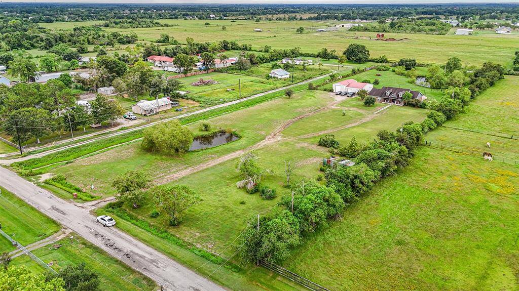 This aerial photo showcases a spacious rural property with expansive green fields and a large home featuring 47 solar panels. The area includes a pond, trees for natural shade, and open land suitable for various outdoor activities. The property is accessible via a road that runs alongside it and is part of a community with neighboring homes in the distance, offering a balance of privacy and community feel.