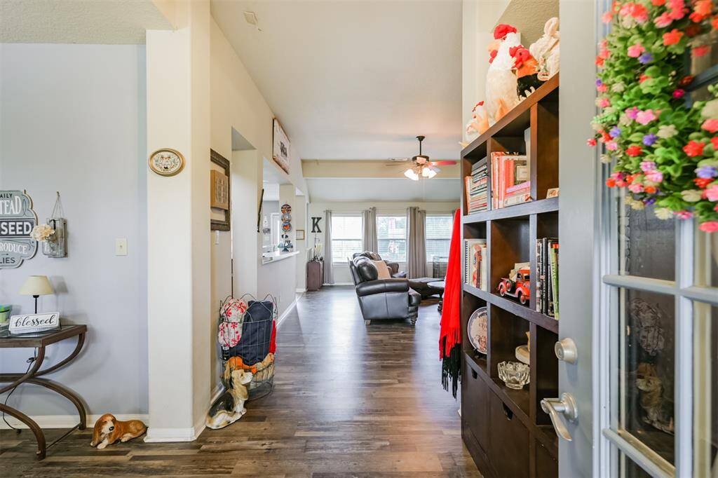 This is a cozy and inviting interior space featuring a warm color palette, with a well-lit hallway leading into a comfortable living area. The space is adorned with personal touches such as wall decorations and a bookshelf filled with various items. The flooring is a beautiful wood laminate, offering a clean and modern look.