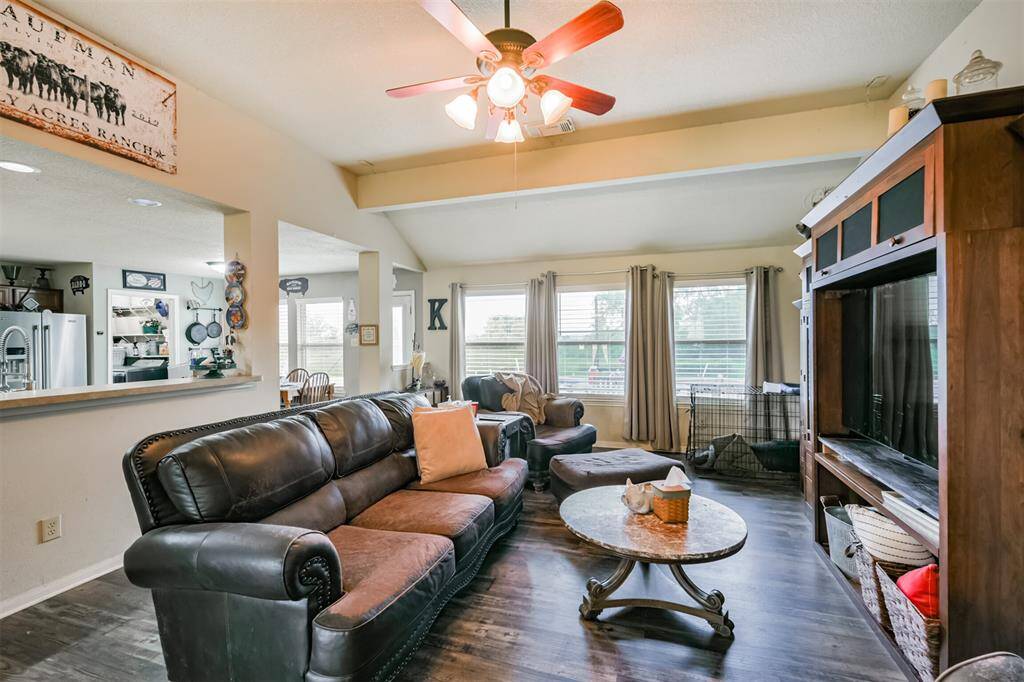 This is a cozy, well-lit living room featuring hardwood floors, and a ceiling fan. The room includes a fireplace with a wooden mantel, large windows allow for plenty of natural light, and the space is open to an adjoining dining area, enhancing the feeling of openness.