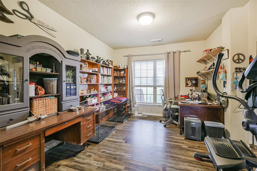 This is a multi-purpose room with wood flooring, currently set up as a home office and exercise area. It features ample natural light from a window with blinds, built-in shelving, and storage cabinetry. The space is versatile and can be reconfigured to suit various needs.