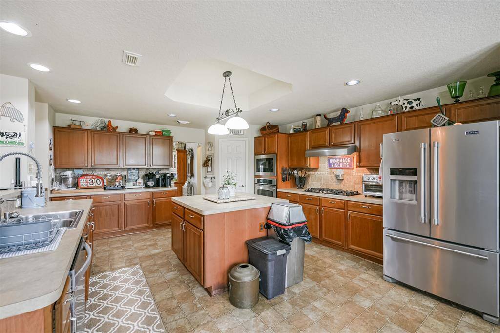 This spacious kitchen features ample cherry wood cabinetry, stainless steel appliances, a central oversized island, and tile flooring, with plenty of natural light.
