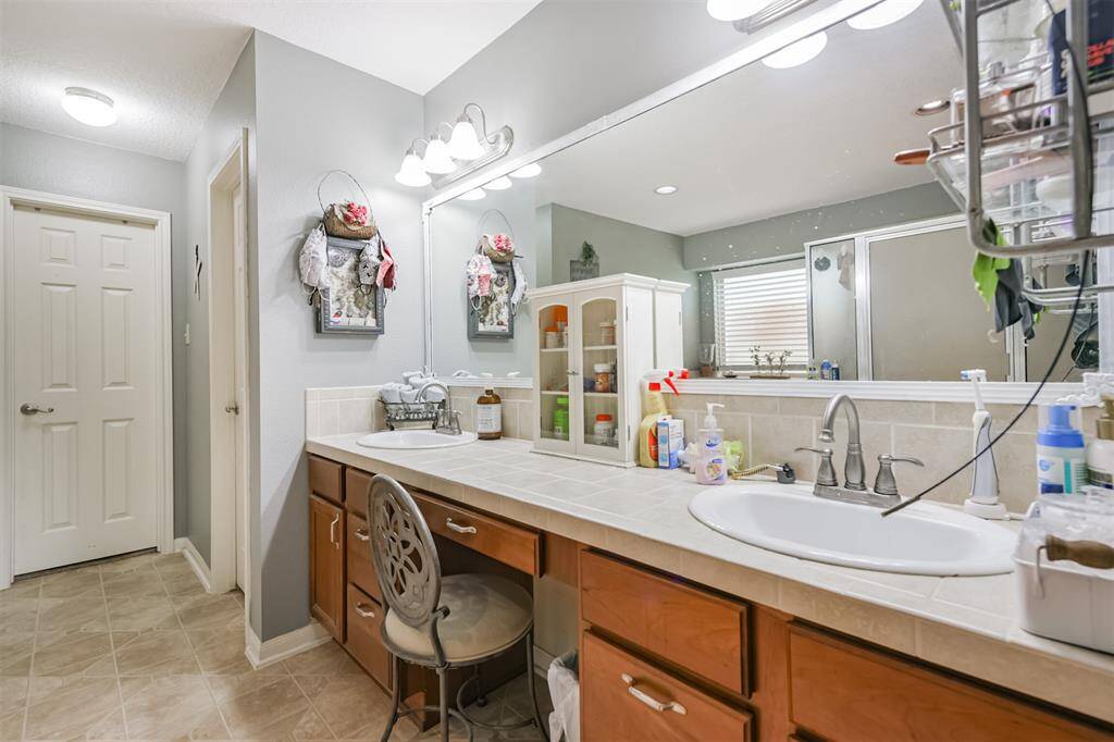 This is a well-lit, spacious bathroom featuring a large vanity with ample counter space and storage, a sink with a modern faucet, a built-in makeup area with a stool, and a full-sized mirror. There is tiled flooring and a neutral color scheme that provides a clean and inviting atmosphere.